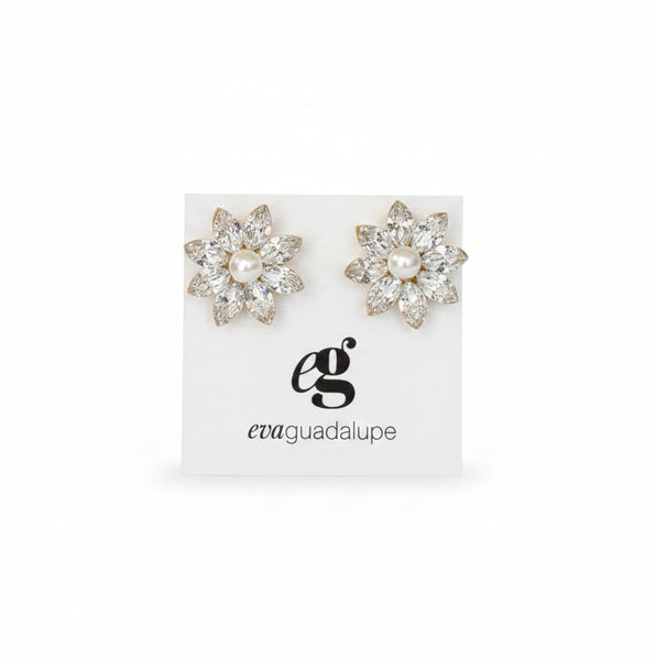 A pair of silver-toned floral stud earrings displayed on a white card inside an open blush pink velvet jewelry box, held by a hand in a busy retail setting.